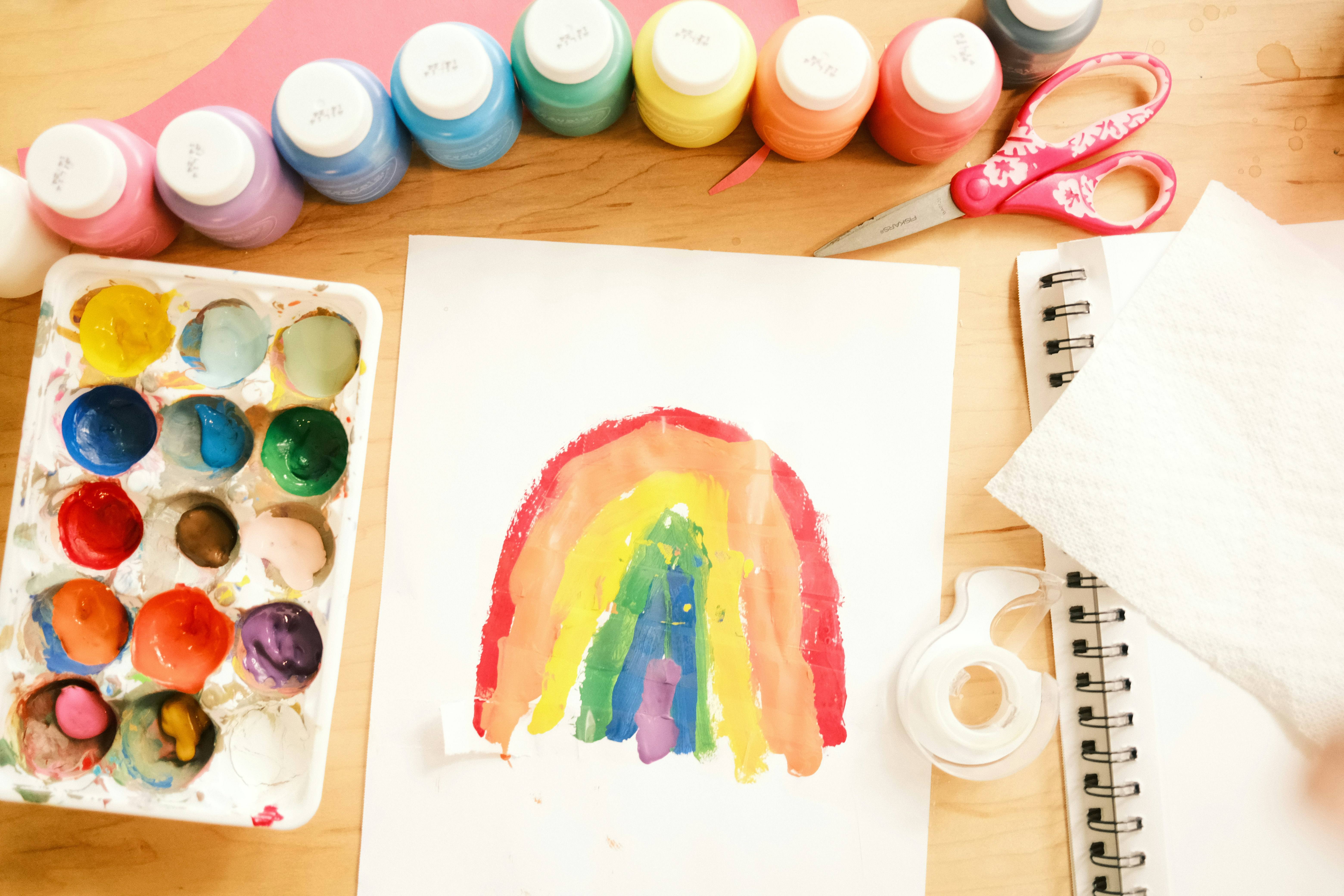 Child's rainbow painting next to paint pots and art supplies